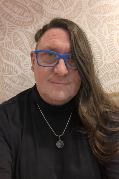 trans woman with long hair and an undercut on the side wearing blue broad rimmed glassed, wearing a silver chain and a black polo neck smiles warmly towards the camera